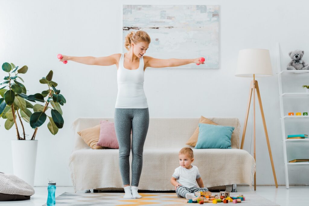Breastfeeding mom doing a low-impact home workout with light dumbbells while her toddler plays nearby